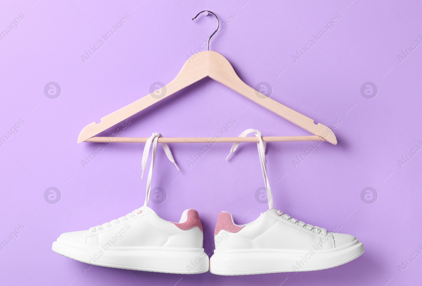 Photo of Stylish sneakers with white shoe laces hanging on wooden hanger against violet background