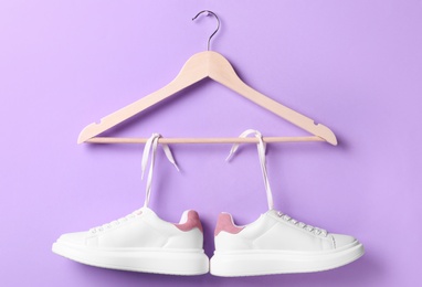 Stylish sneakers with white shoe laces hanging on wooden hanger against violet background