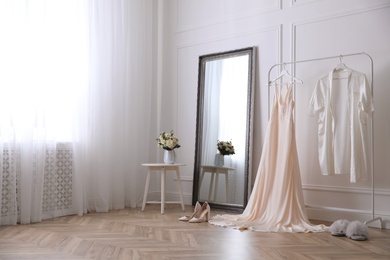 Photo of Elegant wedding dress and silk robe hanging on rack in room