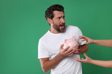 Photo of Scared man trying to protect piggy bank from woman on green background, space for text. Be careful - fraud