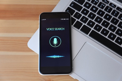 Smartphone with activated voice search app and laptop on wooden table, top view