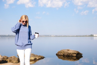 Happy woman in stylish sweater with cup of coffee on beach