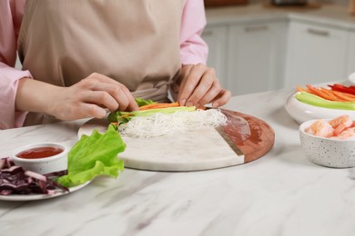 Photo of Making delicious spring rolls. Woman wrapping ingredients into rice paper at white marble table, closeup