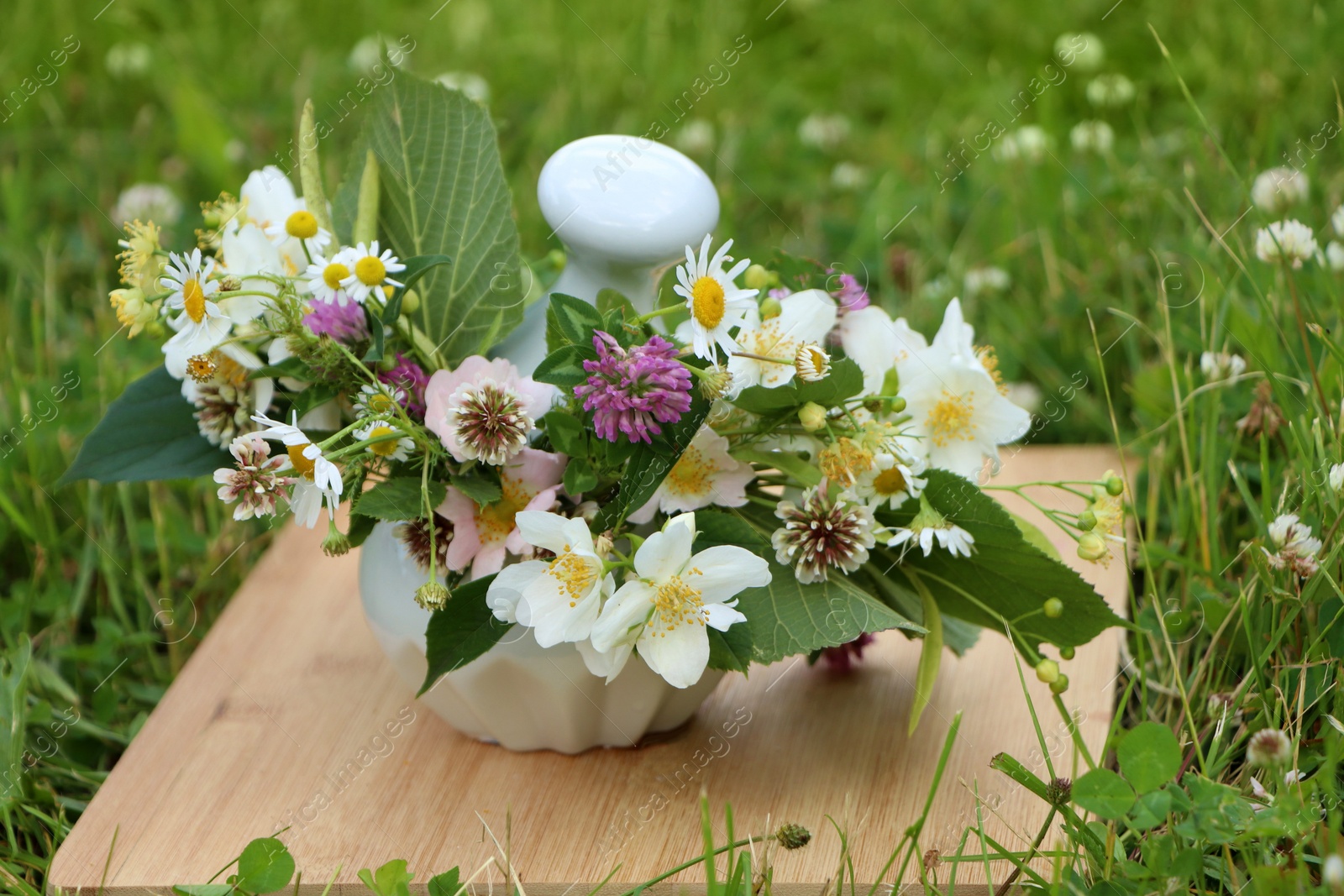 Photo of Ceramic mortar with pestle, different wildflowers and herbs on wooden board in meadow