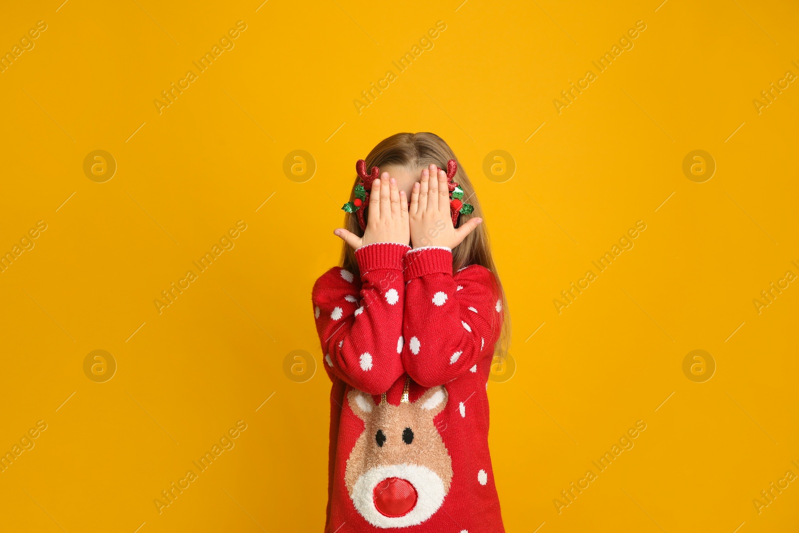 Photo of Cute little girl in Christmas sweater hiding her face on yellow background