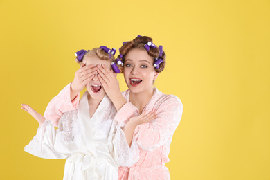 Emotional mother and daughter with curlers on yellow background