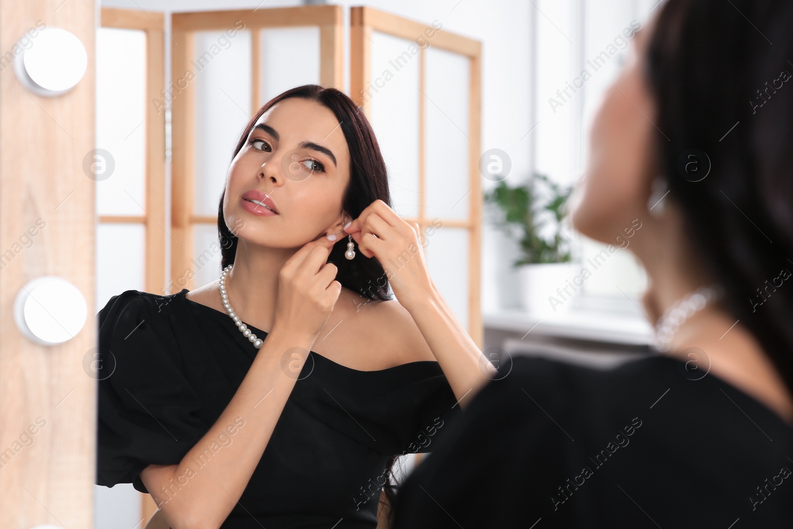 Photo of Young woman trying on elegant pearl earring near mirror indoors