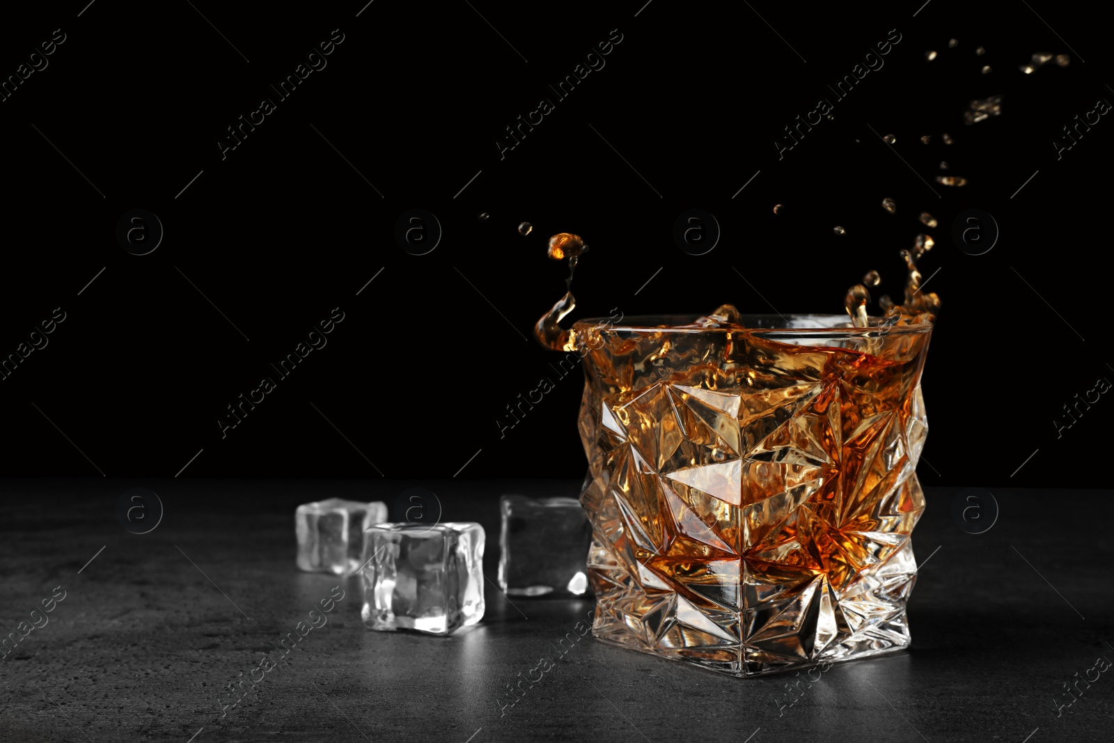 Photo of Splashing golden whiskey in glass with ice cubes on table. Space for text