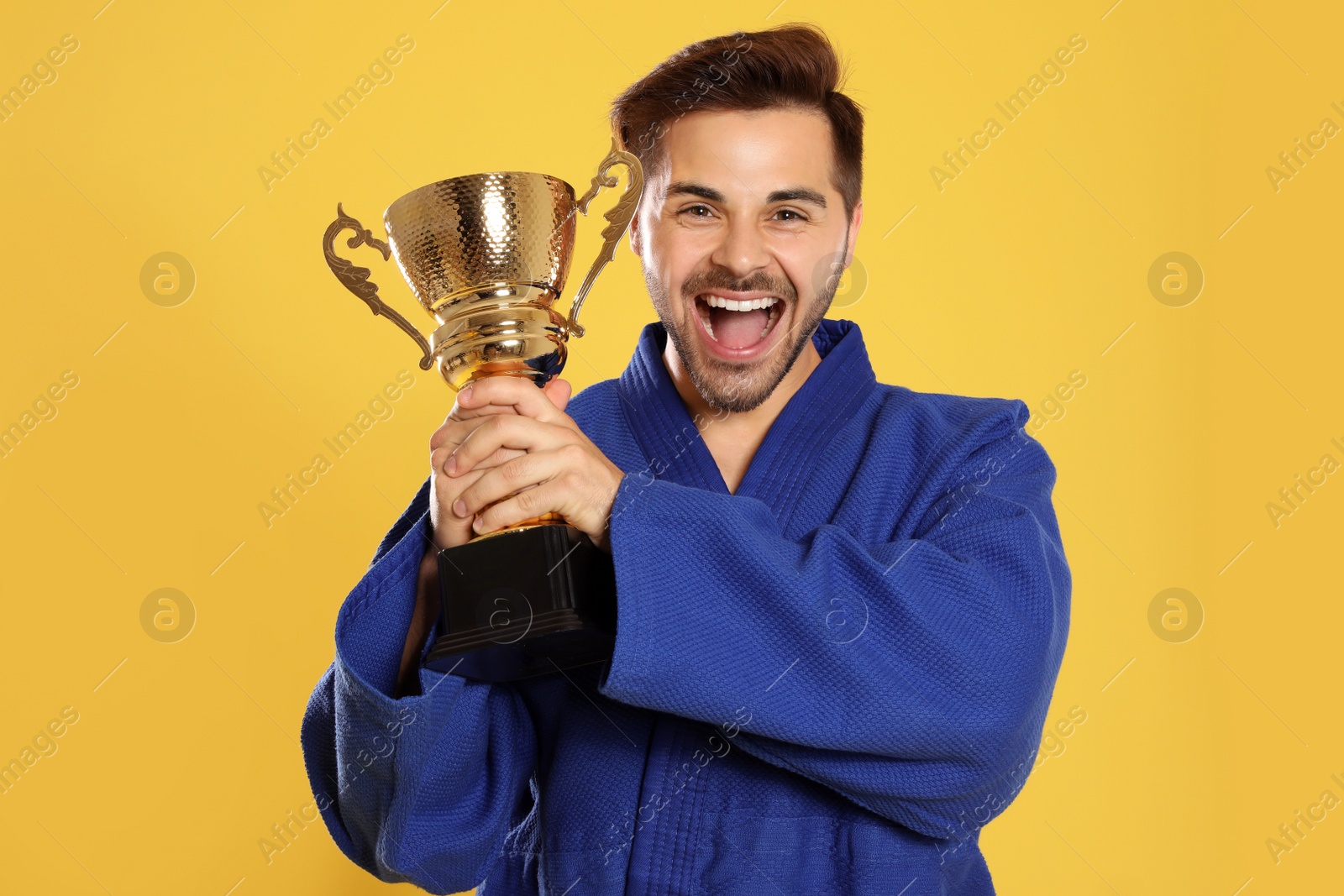 Photo of Portrait of happy young man in blue kimono with gold trophy cup on yellow background