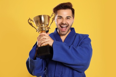Photo of Portrait of happy young man in blue kimono with gold trophy cup on yellow background