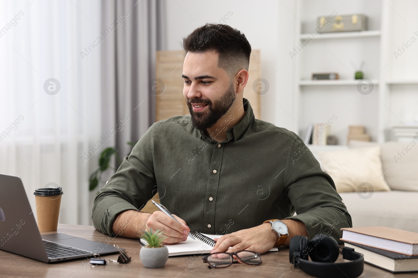 Photo of E-learning. Young man taking notes during online lesson at wooden table indoors