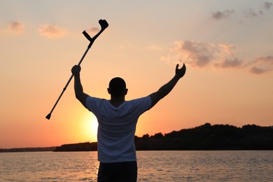 Photo of Man raising elbow crutch up to sky near river at sunset, back view. Healing miracle