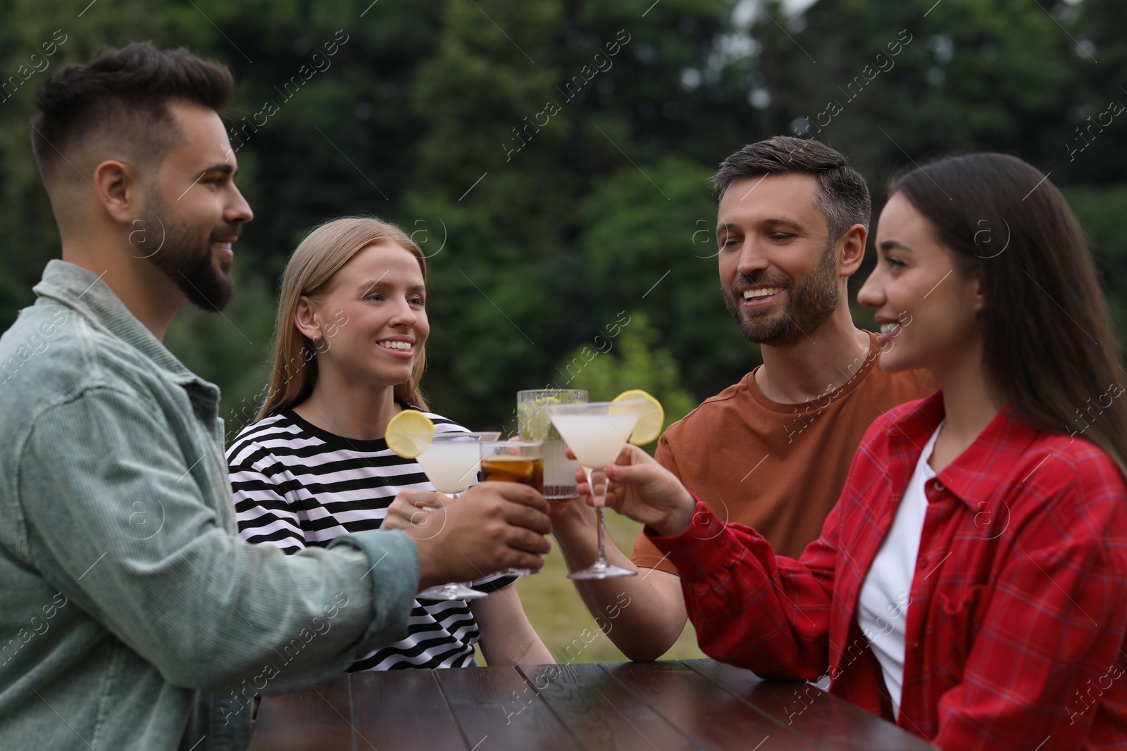 Photo of Happy friends clinking glasses with cocktails at table outdoors