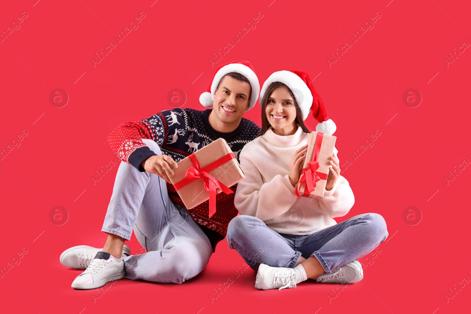 Photo of Beautiful happy couple in Santa hats and sweaters sitting with Christmas gifts on red background