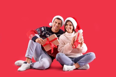Beautiful happy couple in Santa hats and sweaters sitting with Christmas gifts on red background