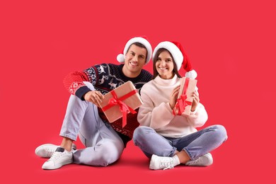 Photo of Beautiful happy couple in Santa hats and sweaters sitting with Christmas gifts on red background