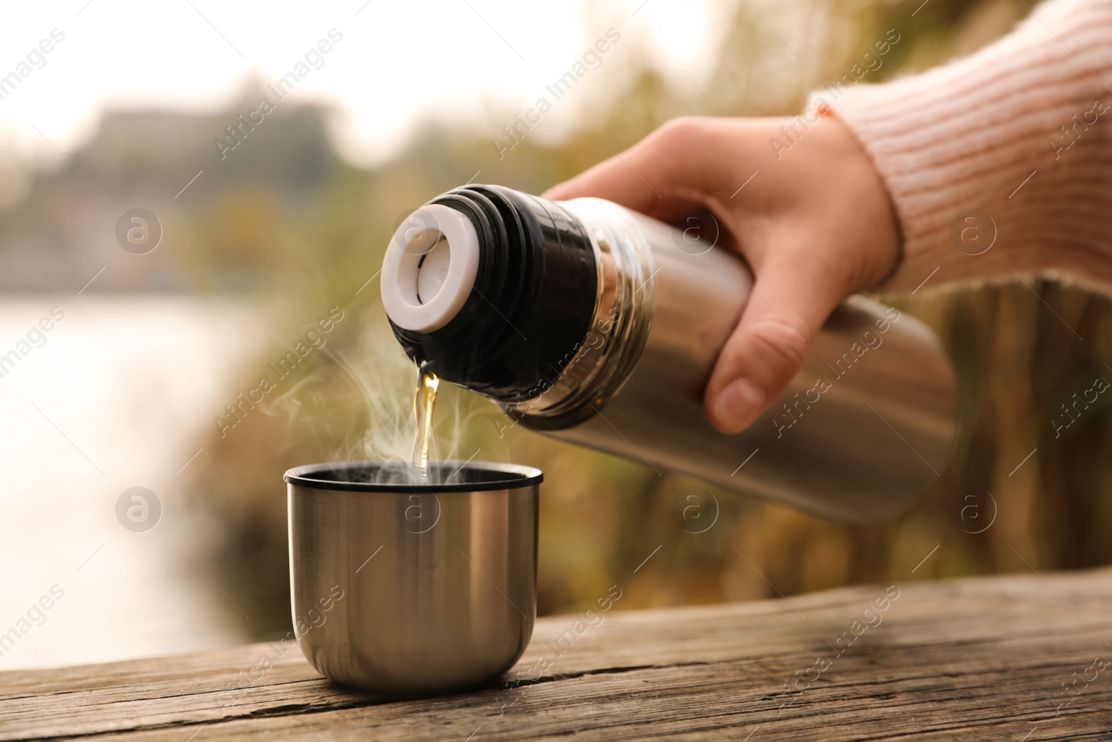 Photo of Woman pouring drink from thermos into cap on wooden pier near river, closeup