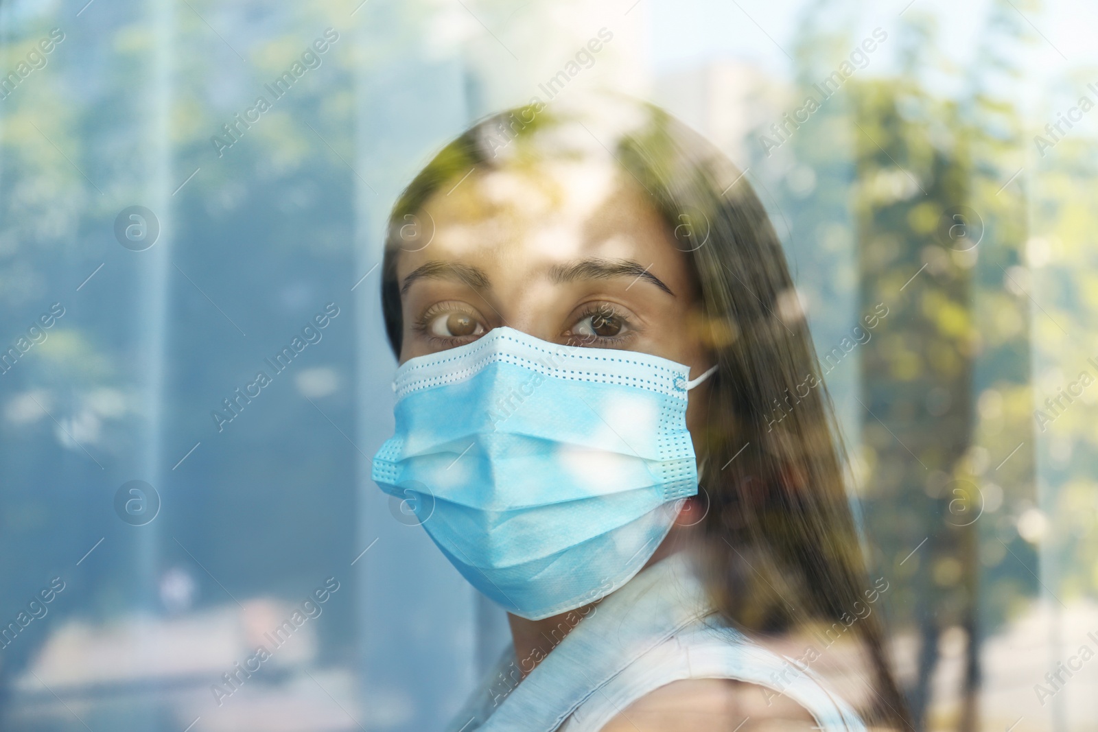 Photo of Stressed woman in protective mask looking out of window, view through glass. Self-isolation during COVID-19 pandemic