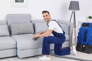 Male worker removing dirt from sofa with professional vacuum cleaner indoors