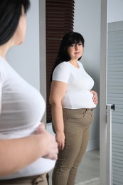 Overweight woman in tight t-shirt and trousers near mirror at home