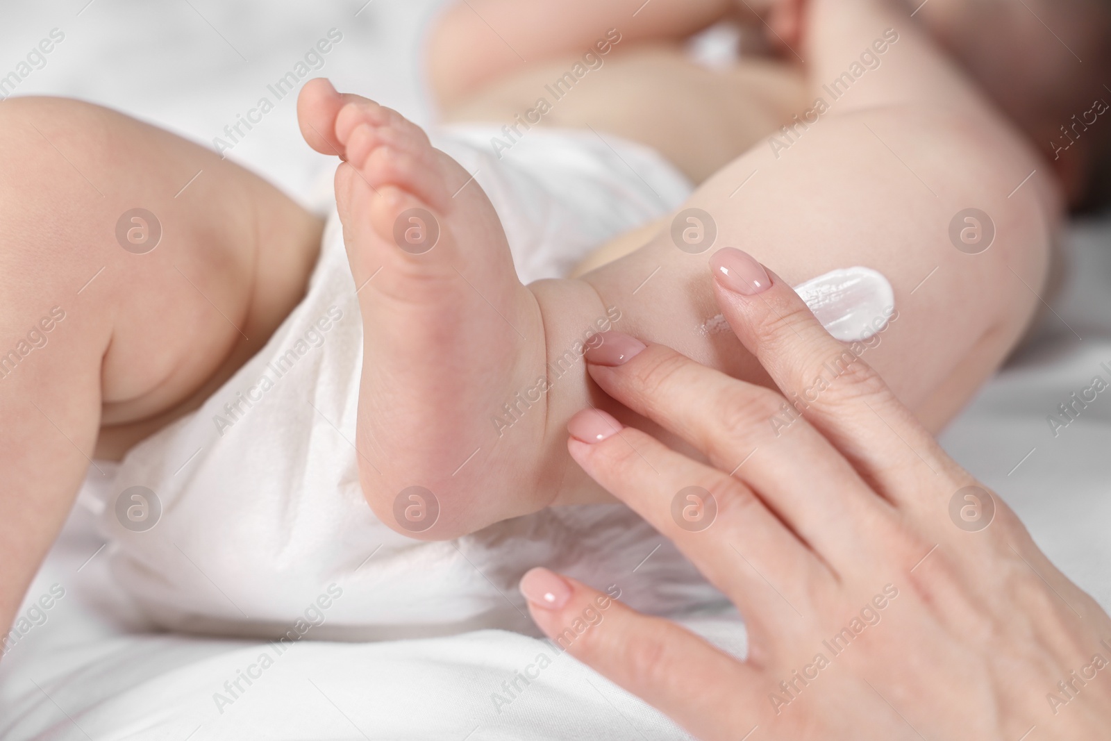 Photo of Woman applying body cream onto baby`s leg on bed, closeup