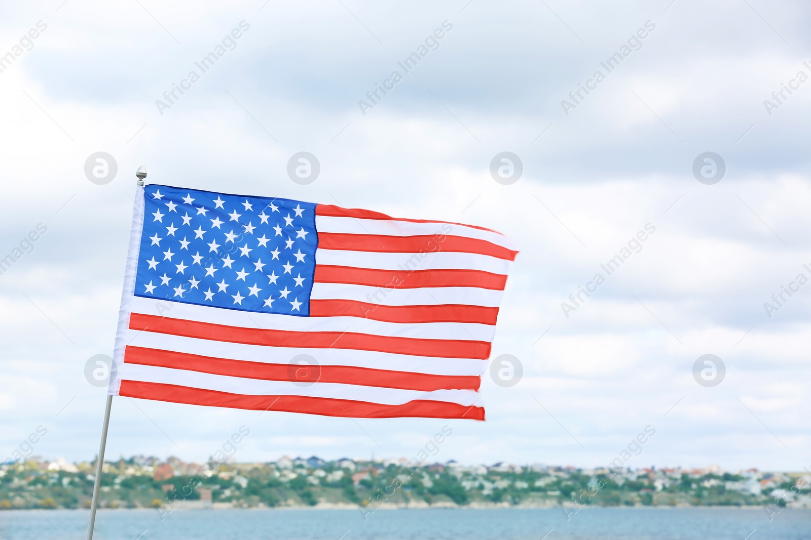 Photo of American flag near river on cloudy day. Space for text