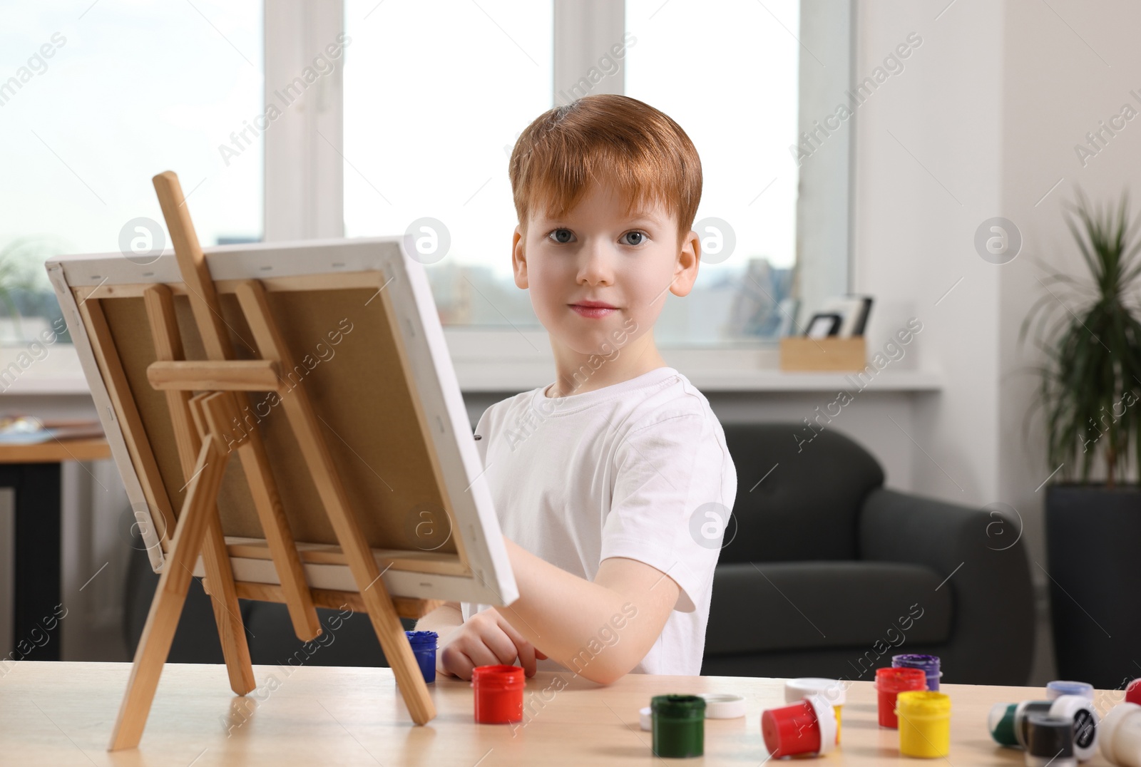 Photo of Little boy painting at table in studio. Using easel to hold canvas