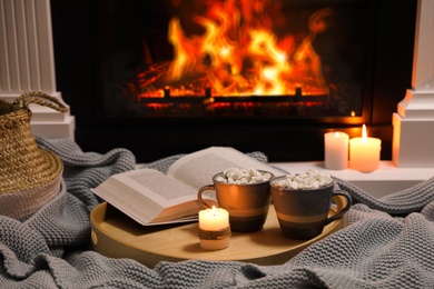 Photo of Cups of hot drink, candle and open book near fireplace indoors