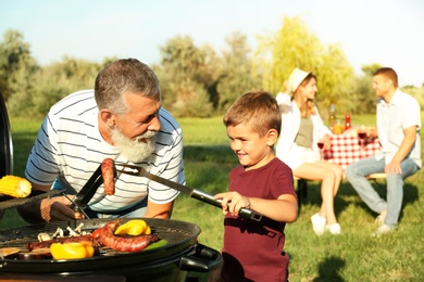 Photo of Grandfather with little boy cooking food on barbecue grill and their family in park