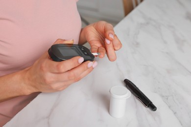 Photo of Diabetes. Woman checking blood sugar level with glucometer at white marble table, closeup