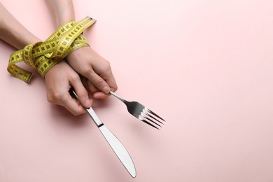 Photo of Diet concept. Woman holding cutlery in hands tied with measuring tape on pink background, top view. Space for text