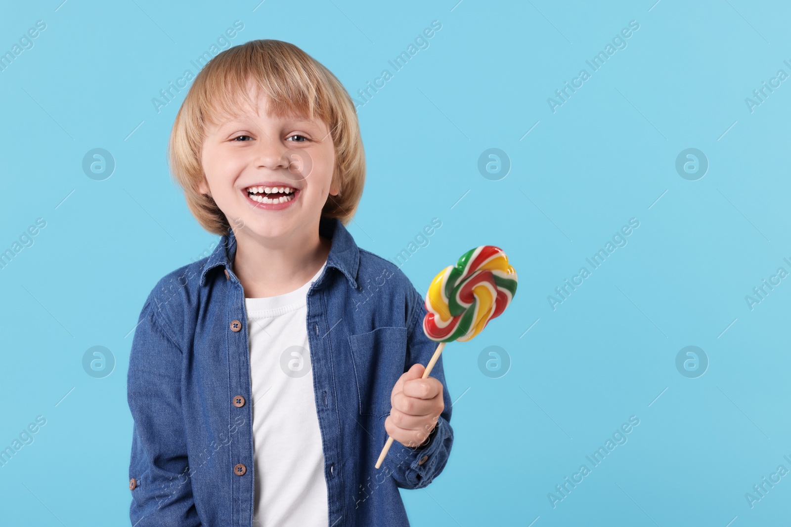 Photo of Happy little boy with colorful lollipop swirl on light blue background, space for text