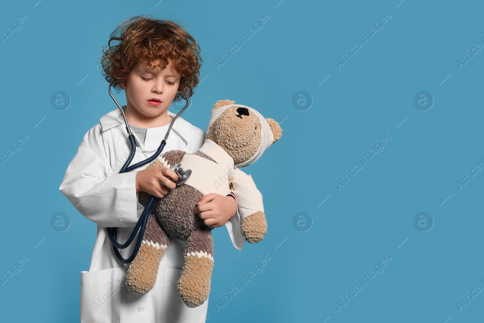 Photo of Little boy in medical uniform examining toy bear with stethoscope on light blue background. Space for text