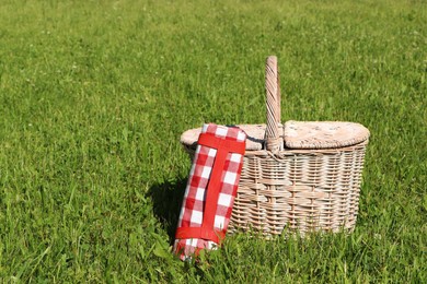 Photo of Rolled checkered tablecloth near picnic basket on green grass outdoors, space for text