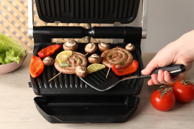 Photo of Woman cooking homemade sausages with bell peppers and mushrooms on electric grill at wooden table, closeup