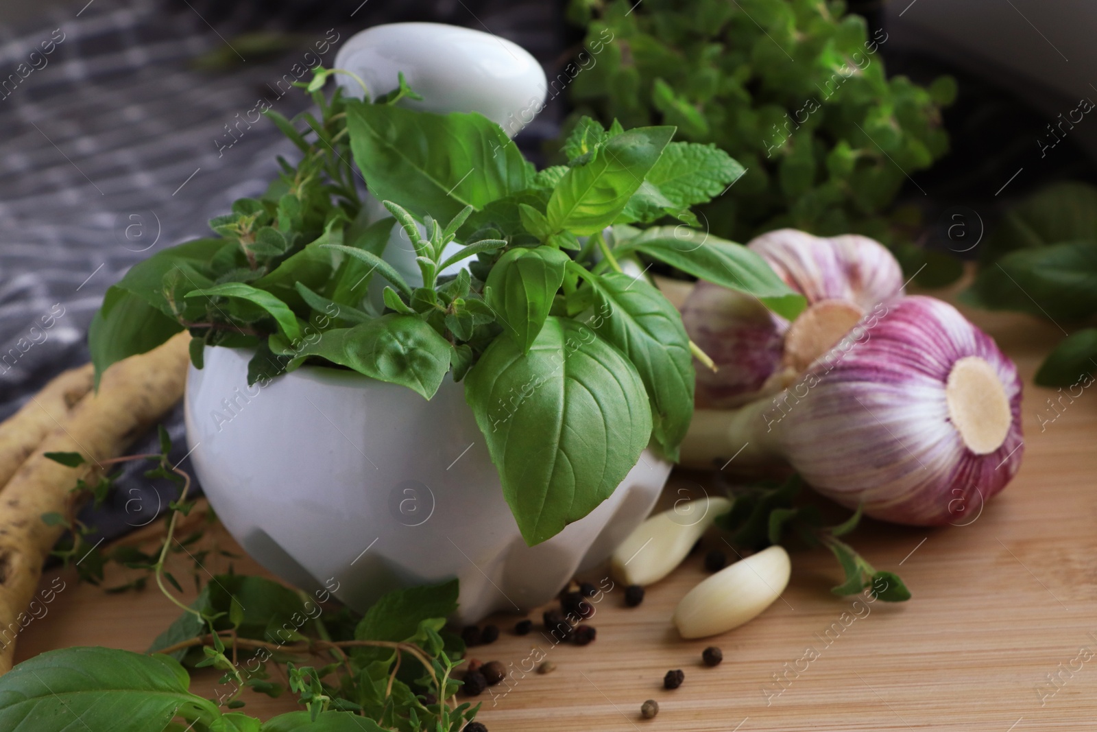 Photo of Mortar with different fresh herbs near garlic, horseradish roots and black peppercorns on wooden table, closeup