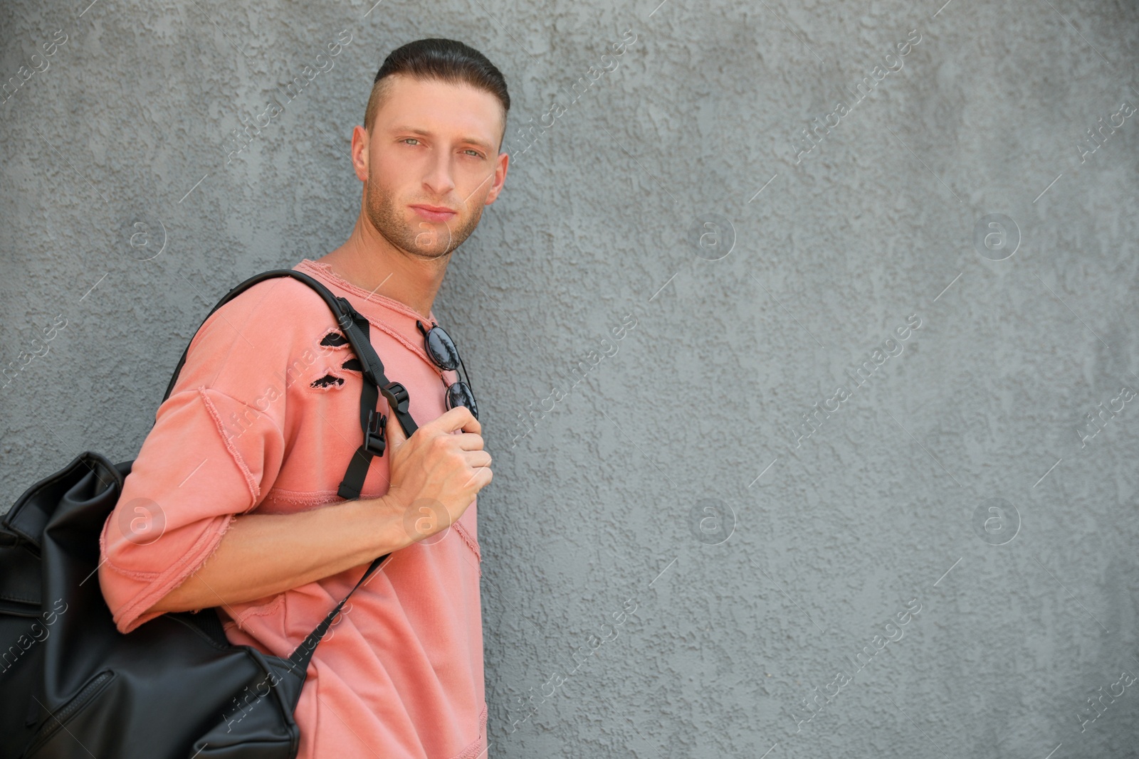 Photo of Handsome young man with stylish sunglasses and bag near grey wall, space for text