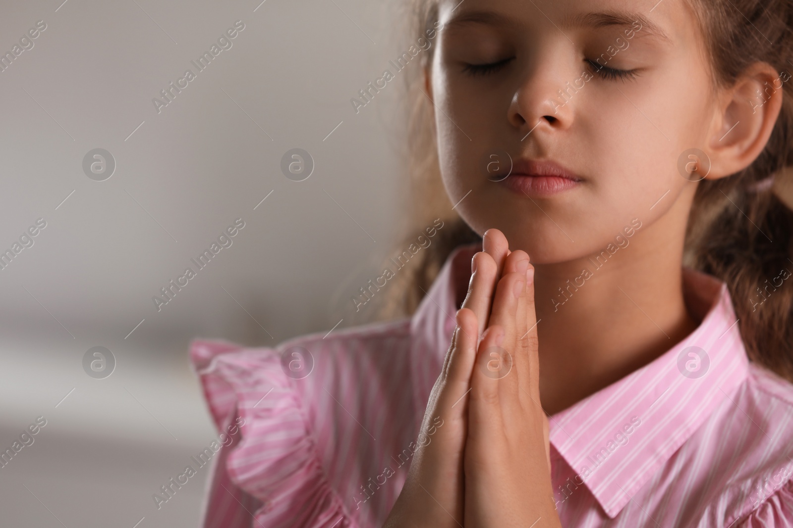 Photo of Cute little girl with hands clasped together praying on blurred background, closeup