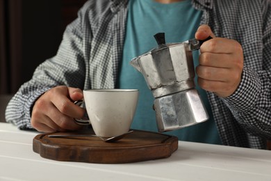 Photo of Man pouring aromatic coffee from moka pot into cup at white wooden table indoors, closeup