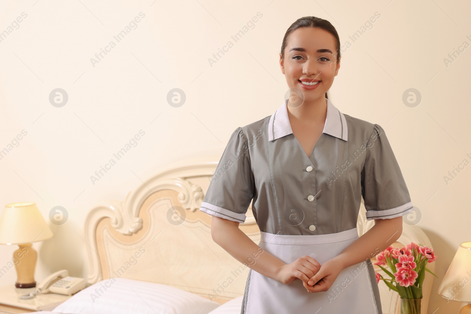 Photo of Portrait of young chambermaid near bed in hotel room. Space for text