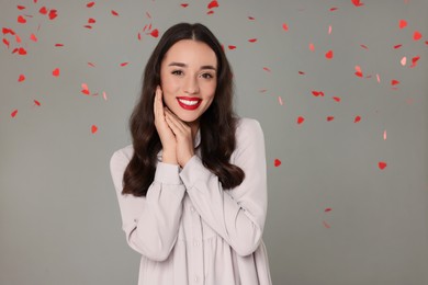 Beautiful young woman under falling heart shaped confetti on grey background