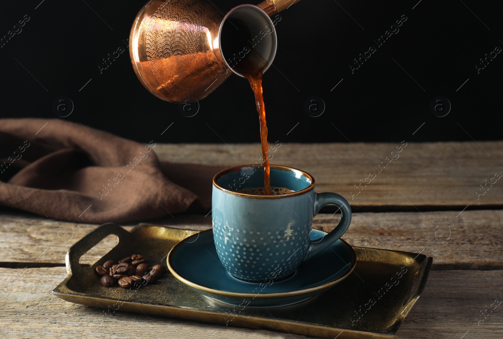 Photo of Turkish coffee. Pouring brewed beverage from cezve into cup at wooden table against black background