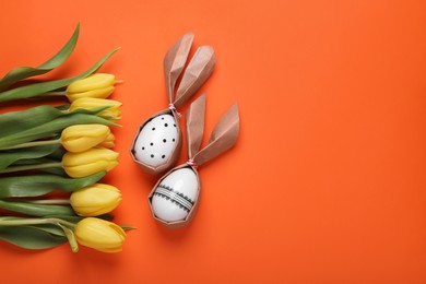 Easter bunnies made of craft paper and eggs near beautiful tulips on orange background, flat lay. Space for text