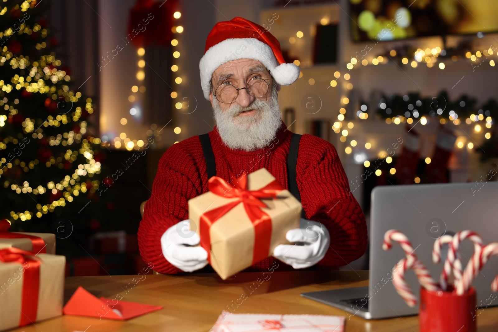 Photo of Santa Claus holding gift box at his workplace in room decorated for Christmas