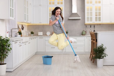 Photo of Enjoying cleaning. Happy woman in headphones jumping with mop in kitchen