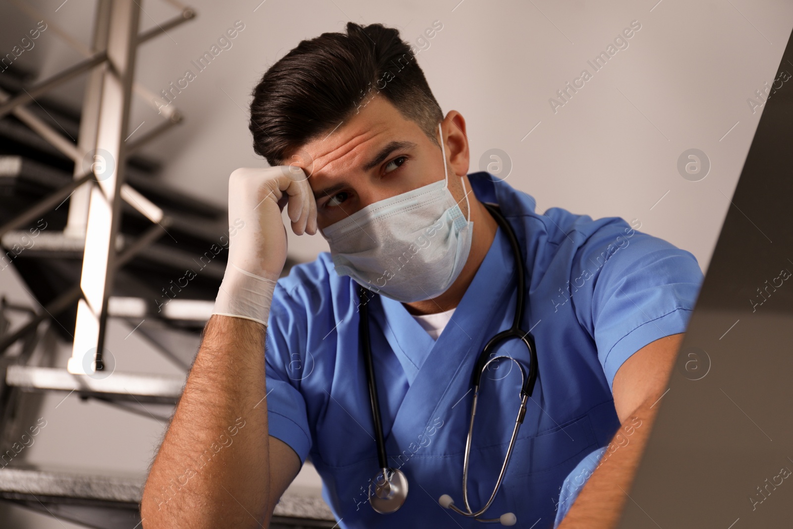 Photo of Sad doctor in facial mask on stairs indoors. Stress of health care workers during coronavirus pandemic