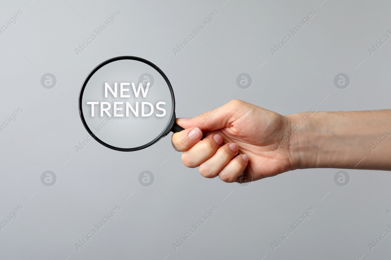 Image of Searching new and popular trends. Woman holding magnifying glass over words on grey background, closeup