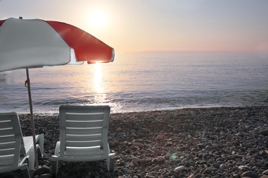 Photo of Empty sunbeds and umbrella on stone beach at sunset