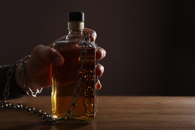 Photo of Alcohol addiction. Man chained with bottle of whiskey at wooden table, closeup. Space for text