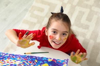 Photo of Little child with painted hands and face at table indoors, view from above